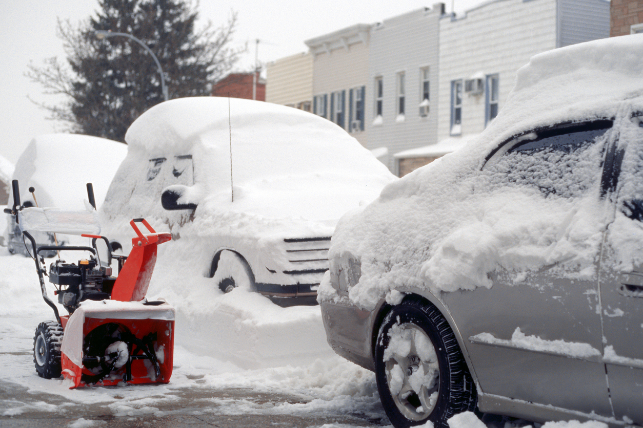 Red snow blower parked on sidewalk next to snow-covered cars.