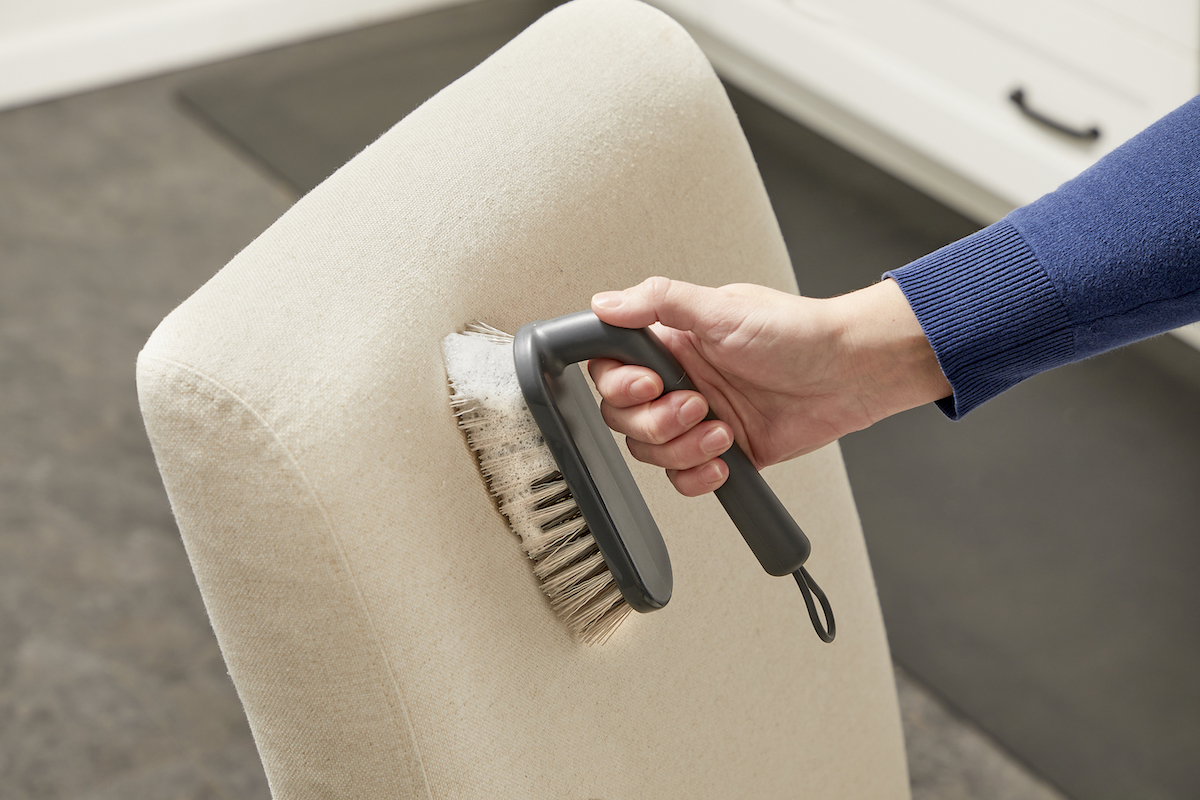 Femme utilisant une brosse à récurer humide pour nettoyer une chaise tapissée beige.