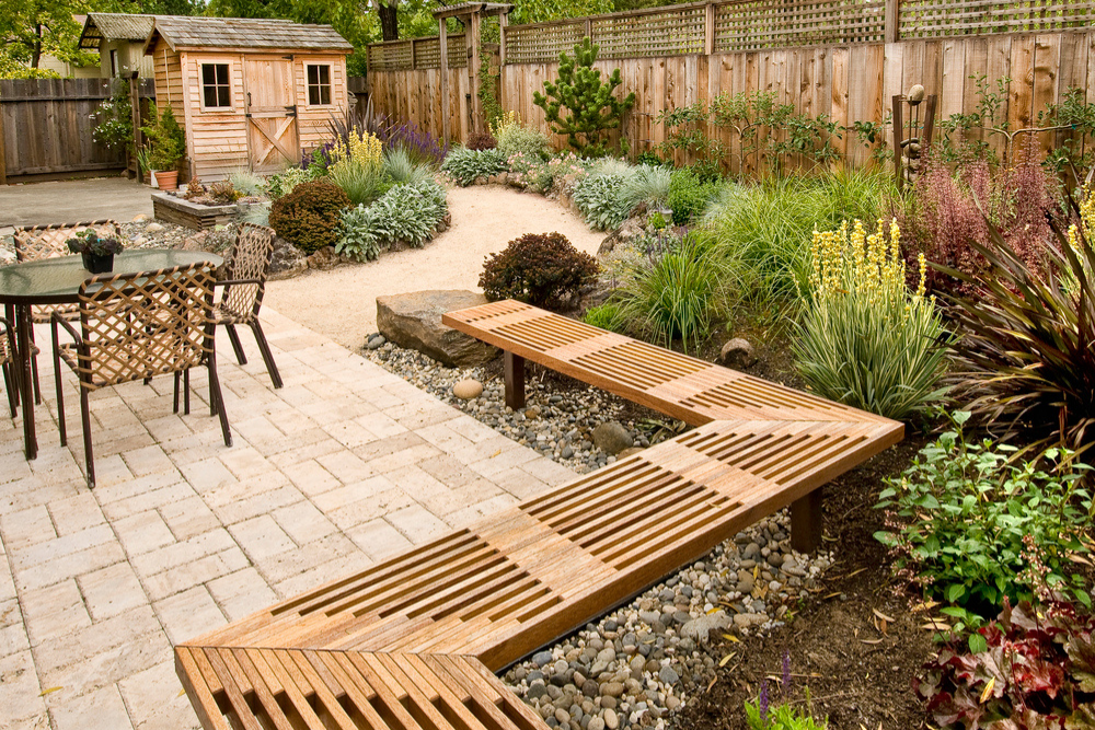 A wooden shed sits in a backyard surrounded by plants with a patio, table, and chairs in the foregound.