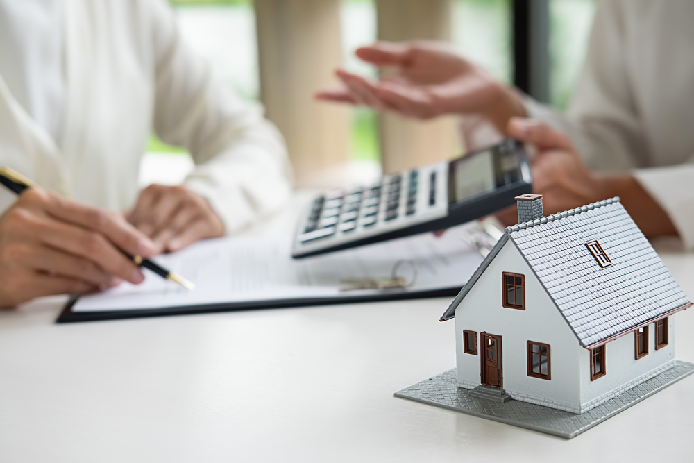 A small white model house sits on a table while two people discuss a document and numbers on a calculator. 