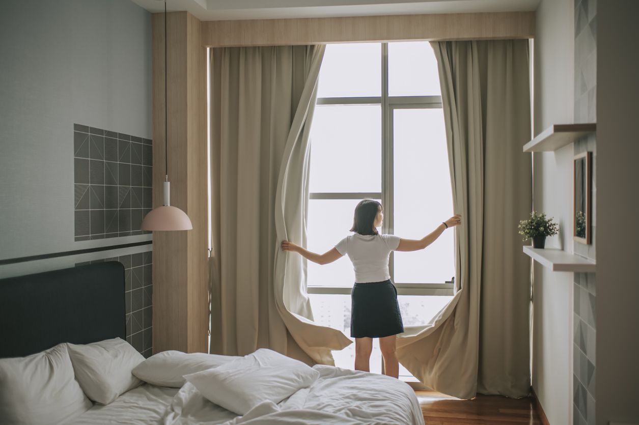 femme ouvrant des rideaux épais dans une chambre à coucher pour laisser entrer la lumière du matin