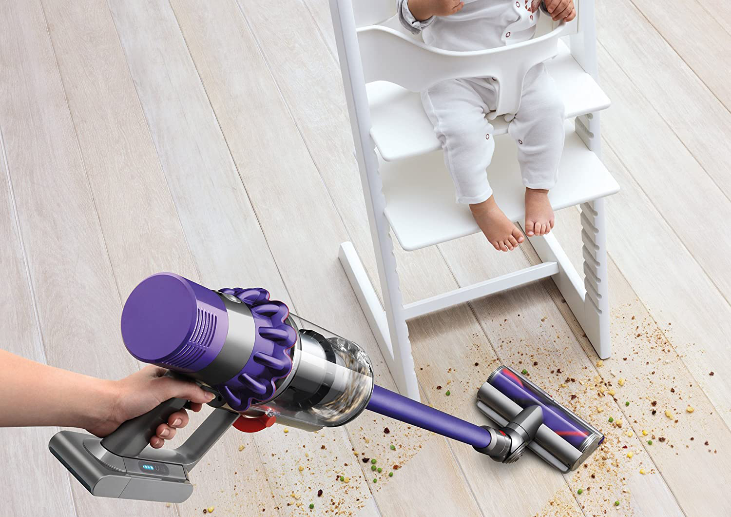 A person cleaning up spilled food on a wood floor under a child in a high chair