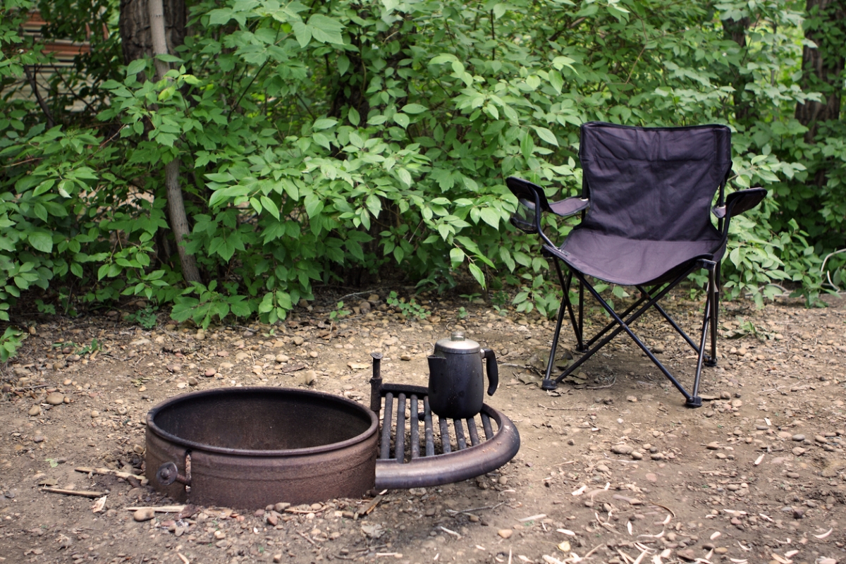 A black camping chair is next to an empty fire pit with a black metal coffee pot sitting on it.