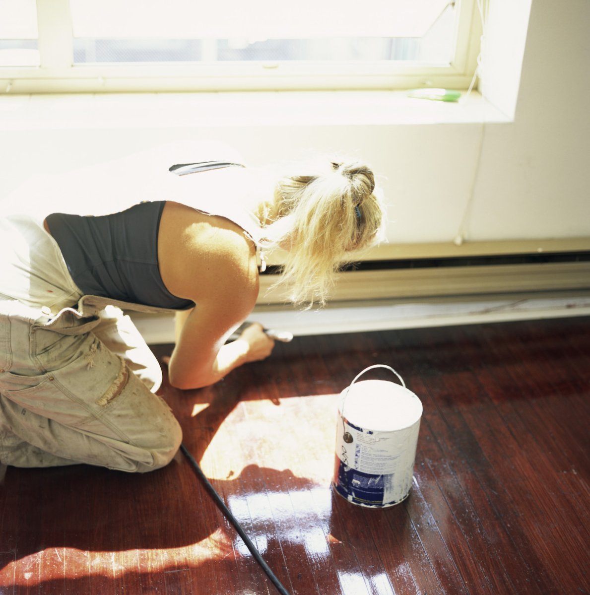 Woman in overalls is crouched on the floor, painting baseboard trim.