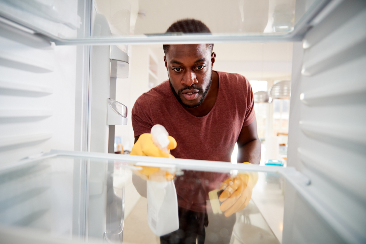 View Looking Out From Inside Empty Refrigerator As Man Wearing Rubber Gloves Cleans Shelves