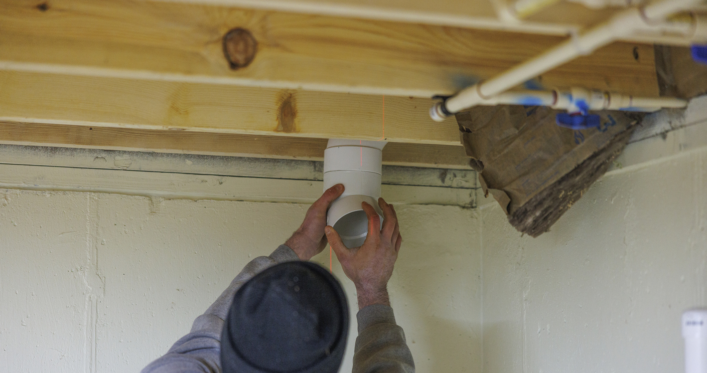 Radon mitigation system installation. A worker is installing a pipe in the basement ceiling.