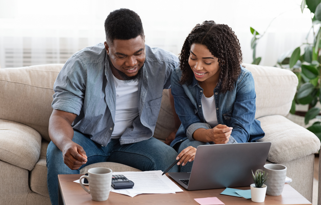 young couple sits on couch in front of window with laptop and financial documents on the coffee table working together