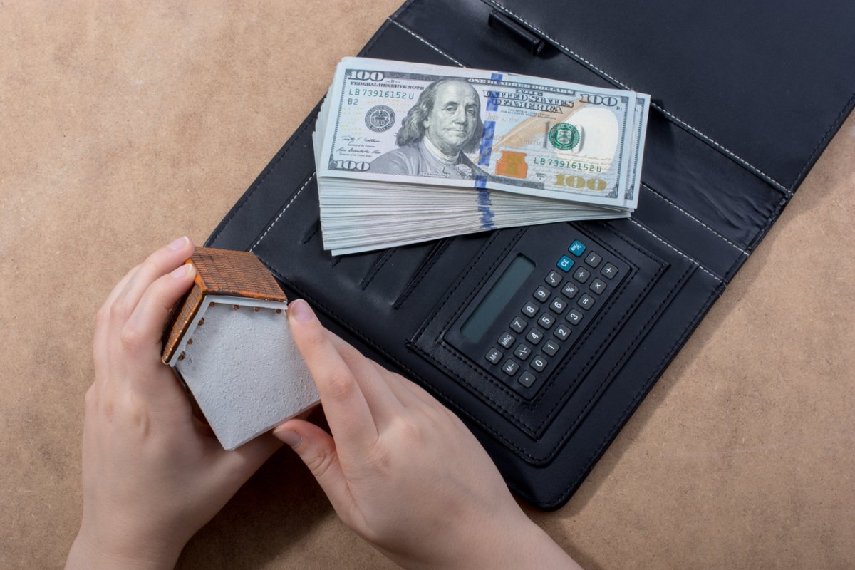 A close up of a person's hands holding a small model house next to a calculator and a pile of cash.