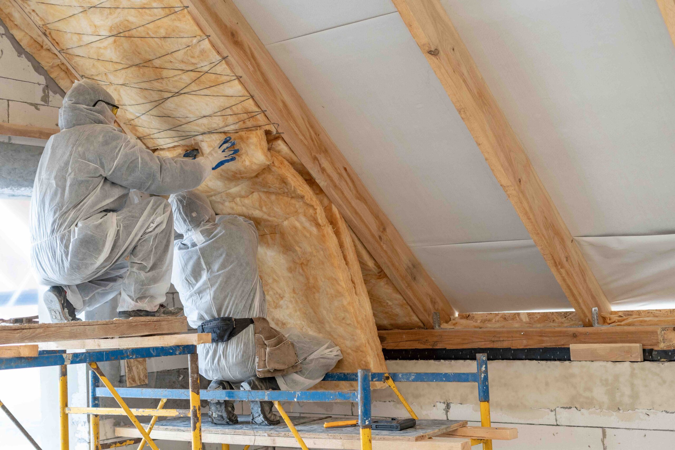 Vue arrière de deux ouvriers professionnels en salopette travaillant ensemble avec une isolation en laine de roche, debout à l'intérieur d'une nouvelle maison en construction, fixant le matériau de chaleur au plafond et au mur avec espace de copie