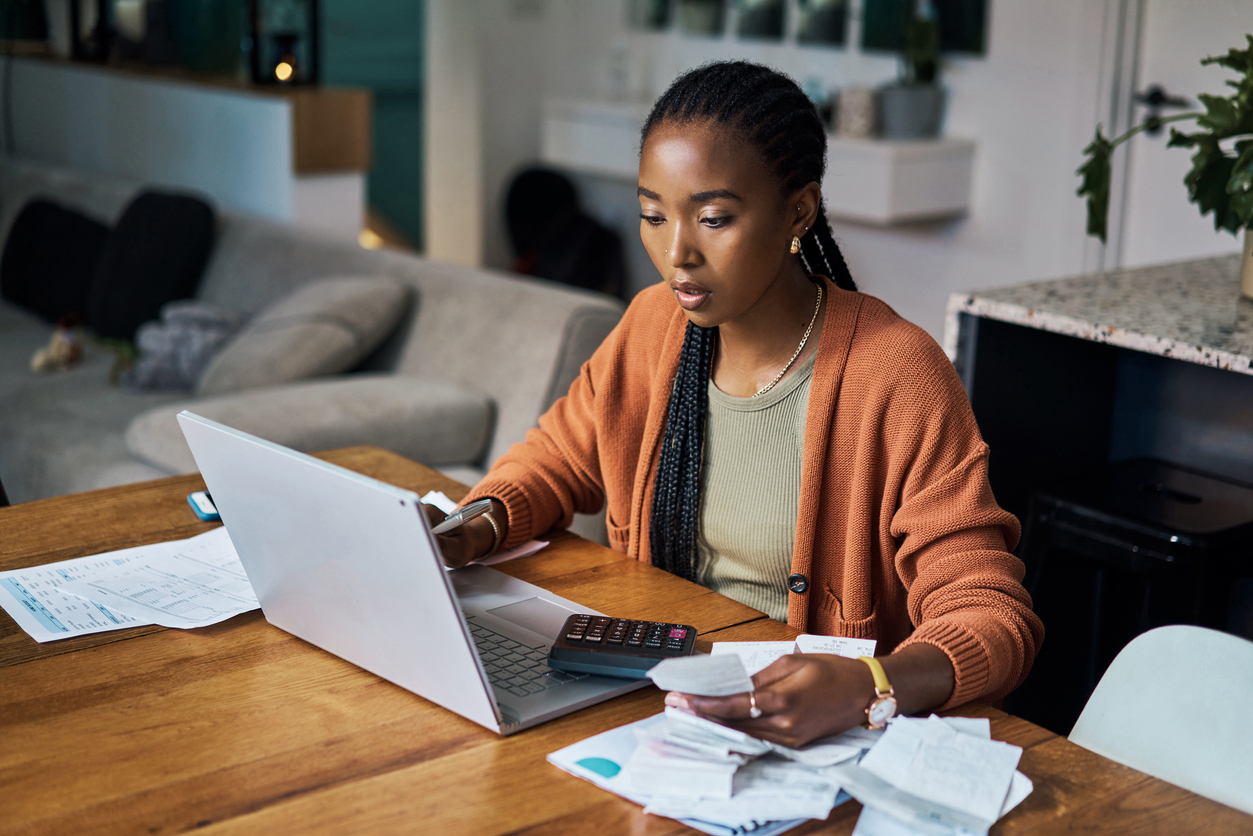 Young woman in orange sweater does taxes with laptop on kitchen table.