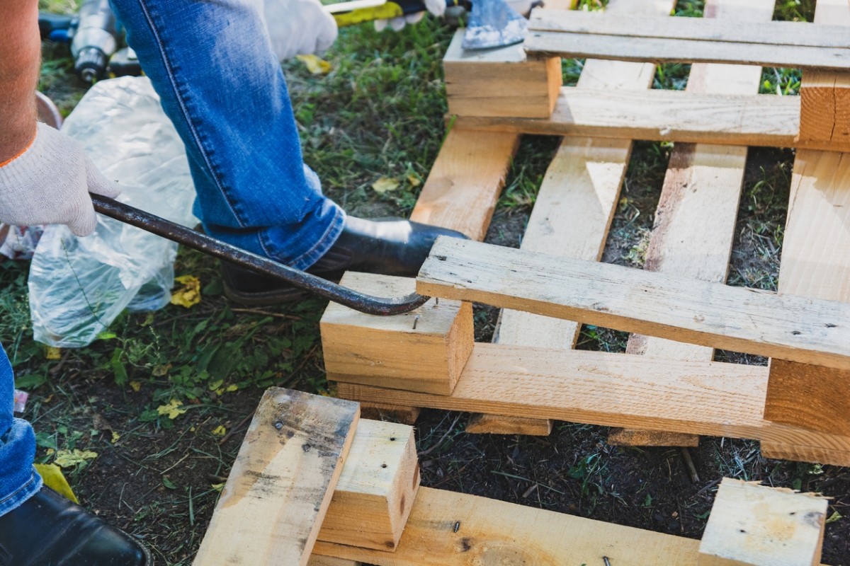 A person is breaking a wooden pallet by using a crowbar and their foot.