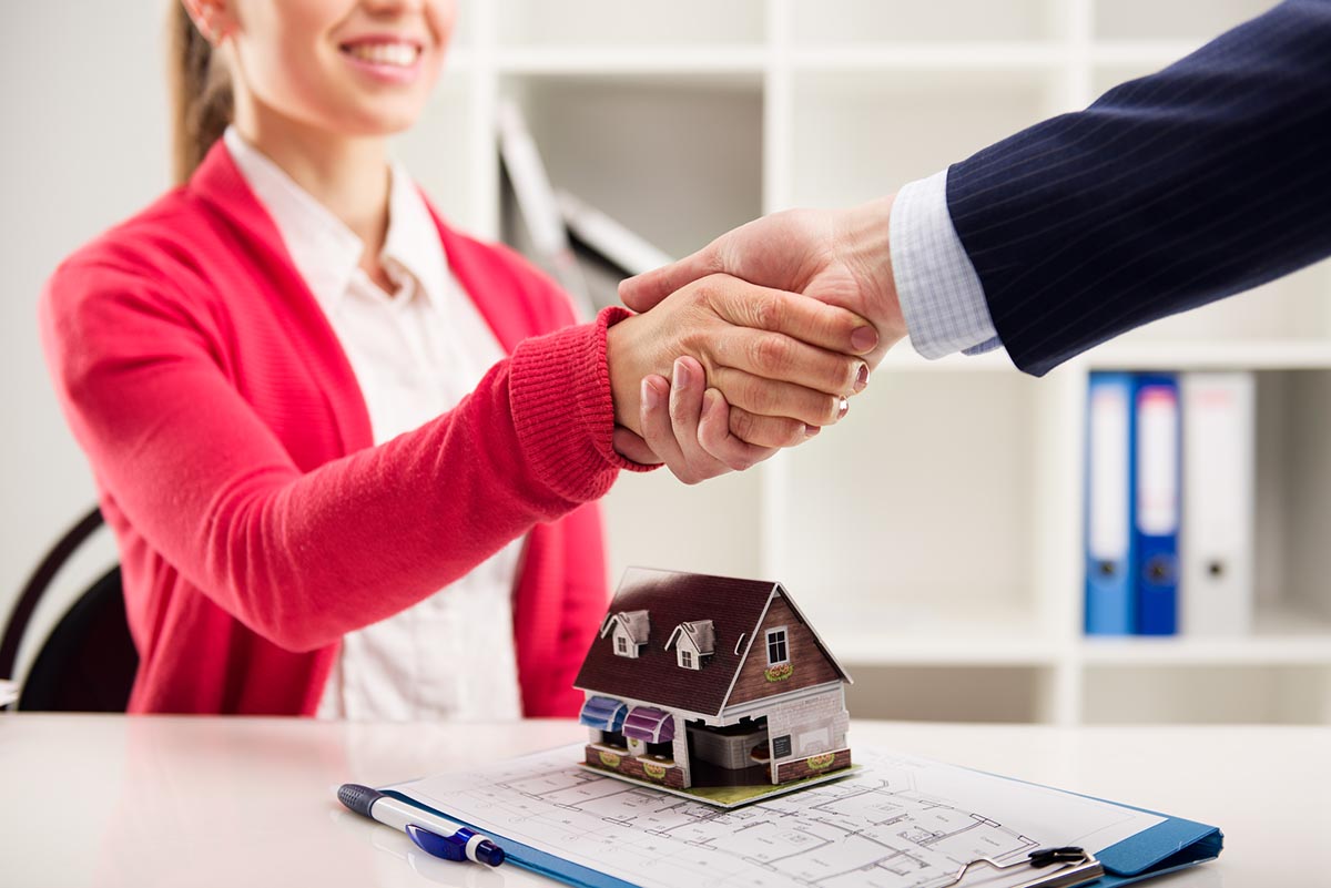 Two people shake hands over a small model house sitting atop a floor plan.