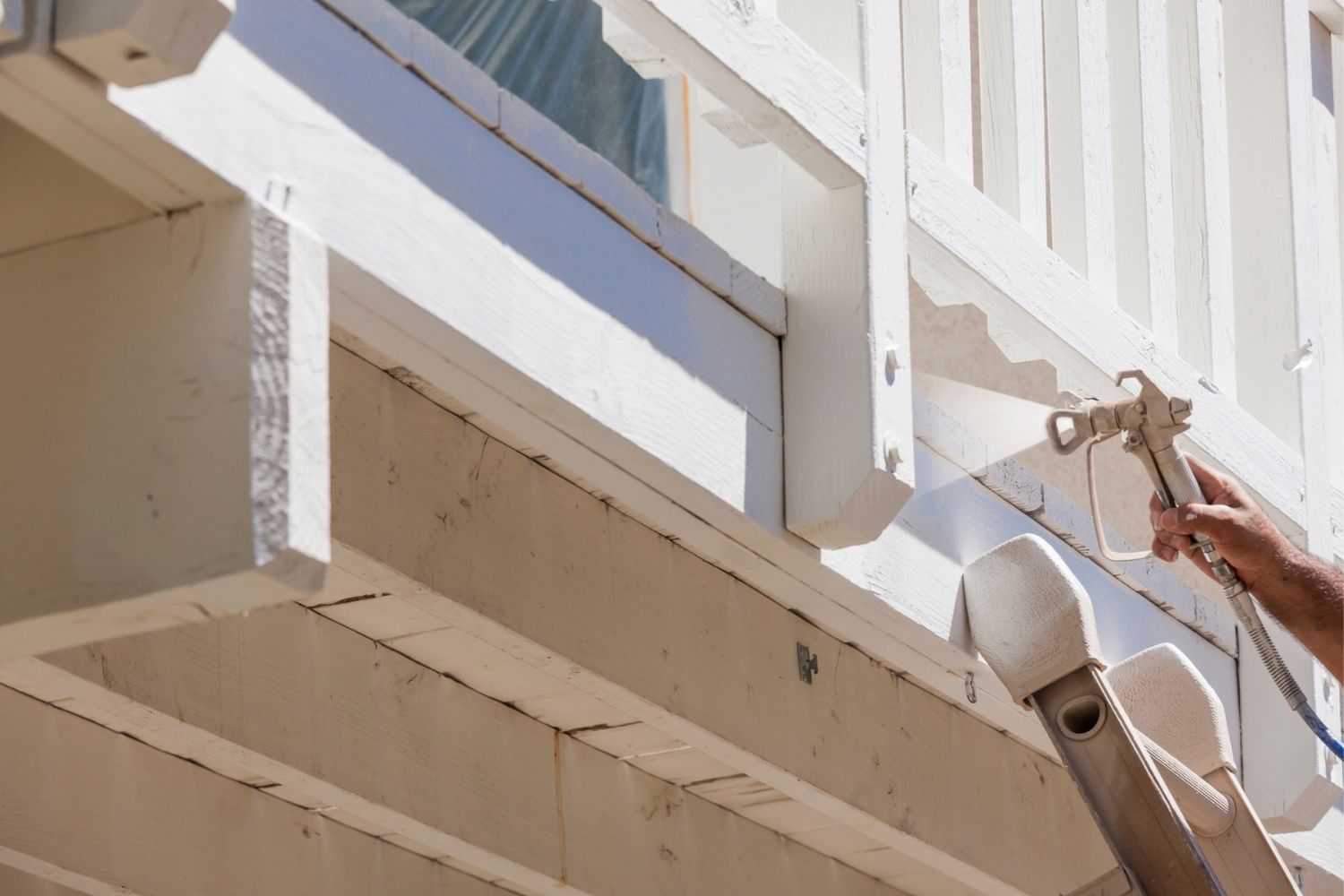 A close-up of a professional using a paint sprayer to apply white paint to a wooden deck.