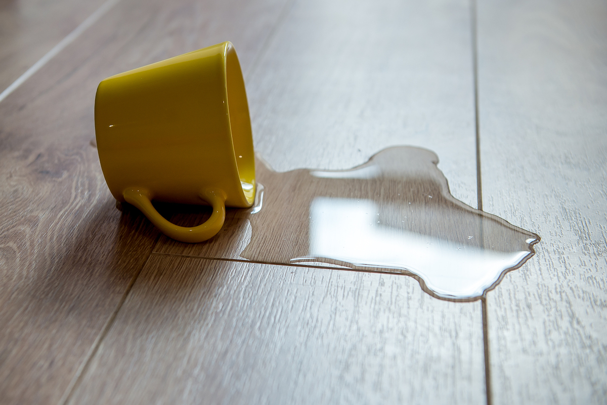 A yellow cup is tipped over with water spilling out onto a wooden laminate floor.