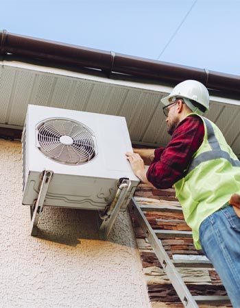 A worker in a hard hat and a safety vest climbs a ladder to repair an AC unit. 