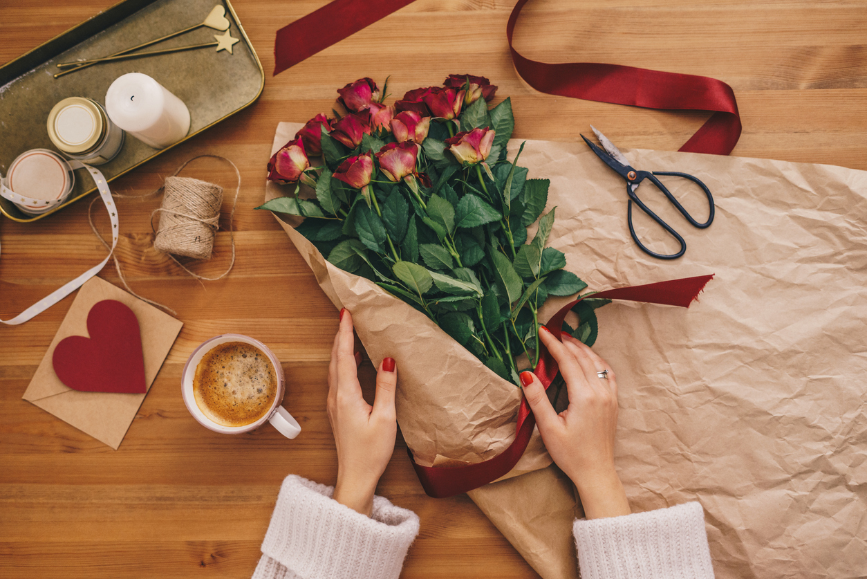 Top view of woman's hands creating a bouquet of rose flowers on a wooden table with ribbons and paper