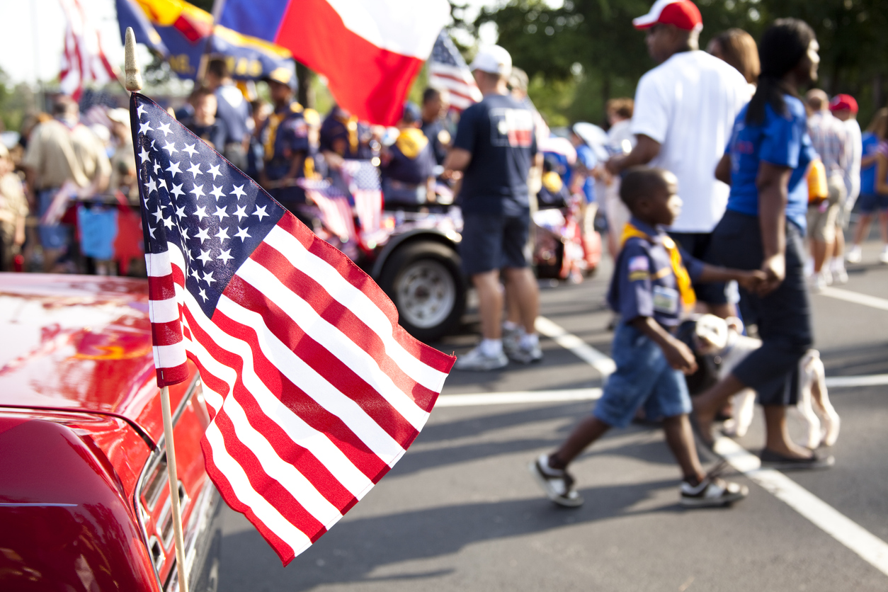 American flag flying from the back of a vintage red car in the foreground of a block party in a parking lot