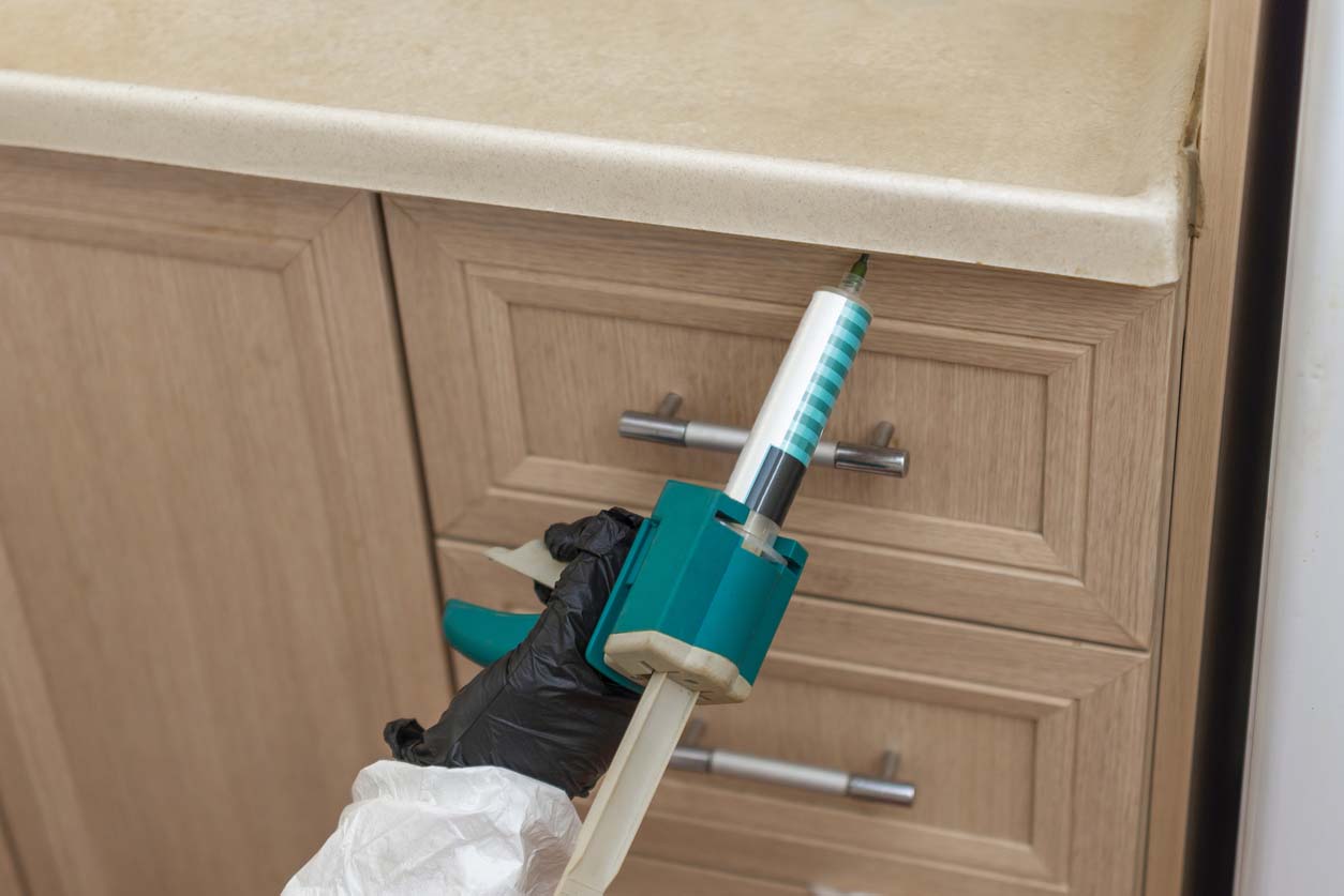 A person wearing personal protective equipment, including a latex glove, applies a pest control product to a kitchen cabinet.