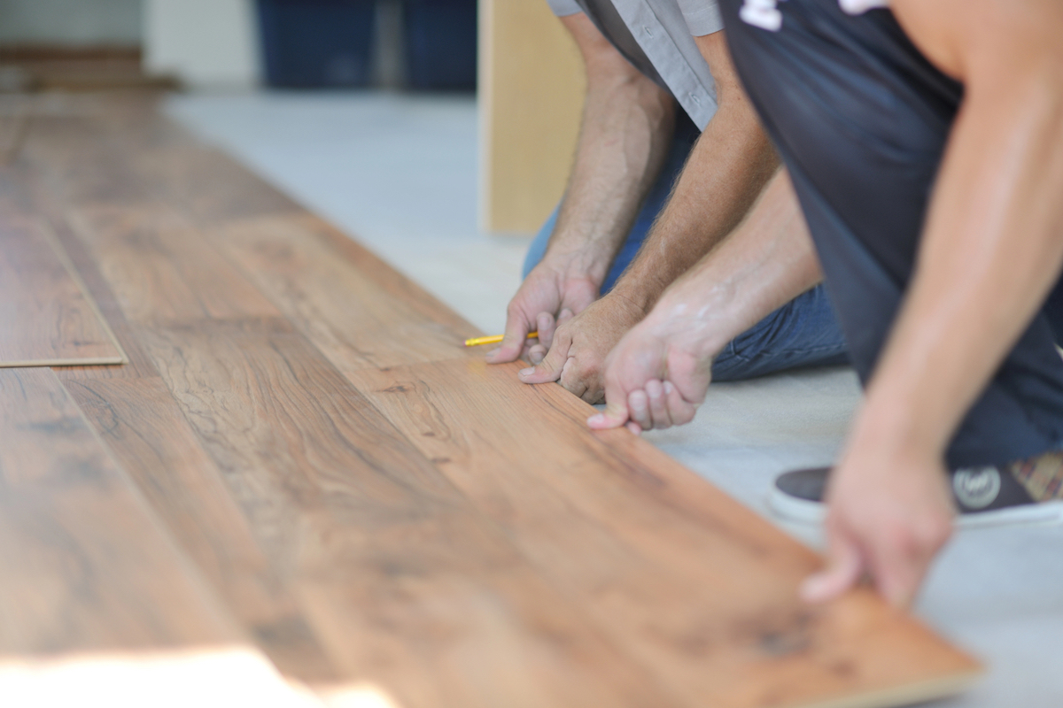 Two men are kneeling on the floor while installing wooden laminate flooring.