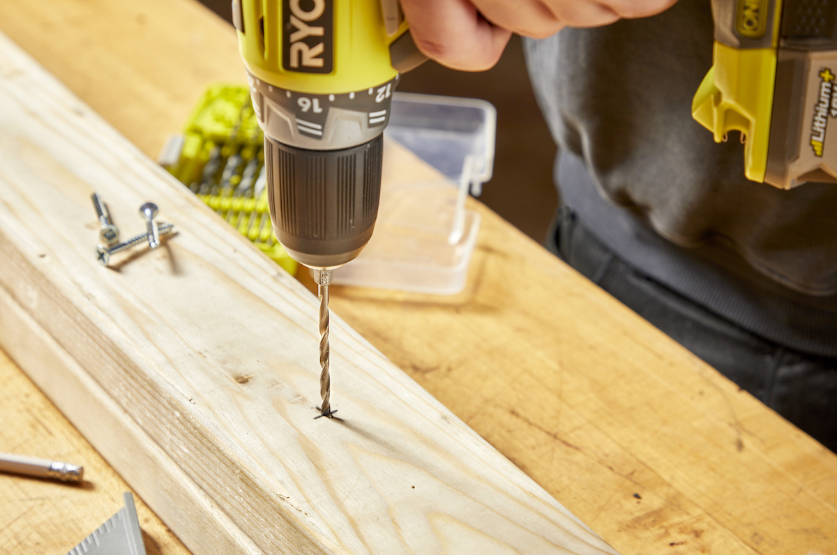 Man points drill bit into an X on a wood plank.