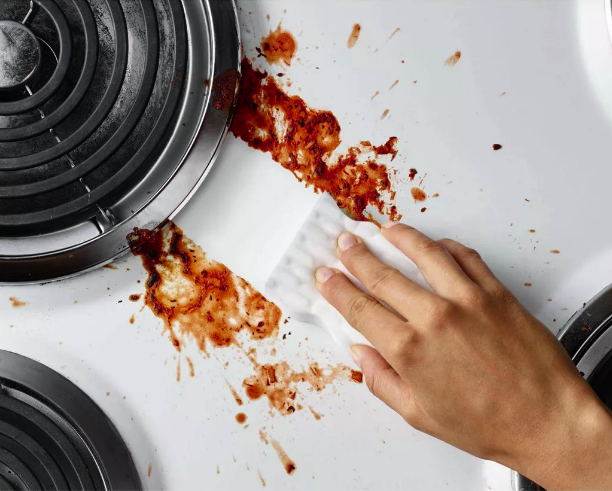 stovetop with heavy dry stain from red sauce being cleaned with a magic eraser that leaves a clean white streak