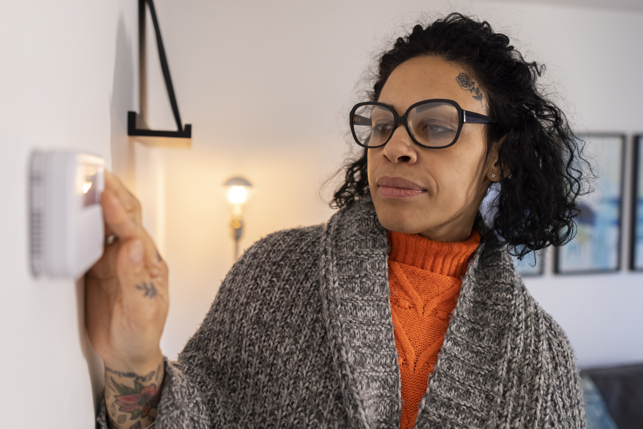 Woman in her 40s with glasses adjusting her home's central heating thermostat. She is wearing a blanket around her shoulders.