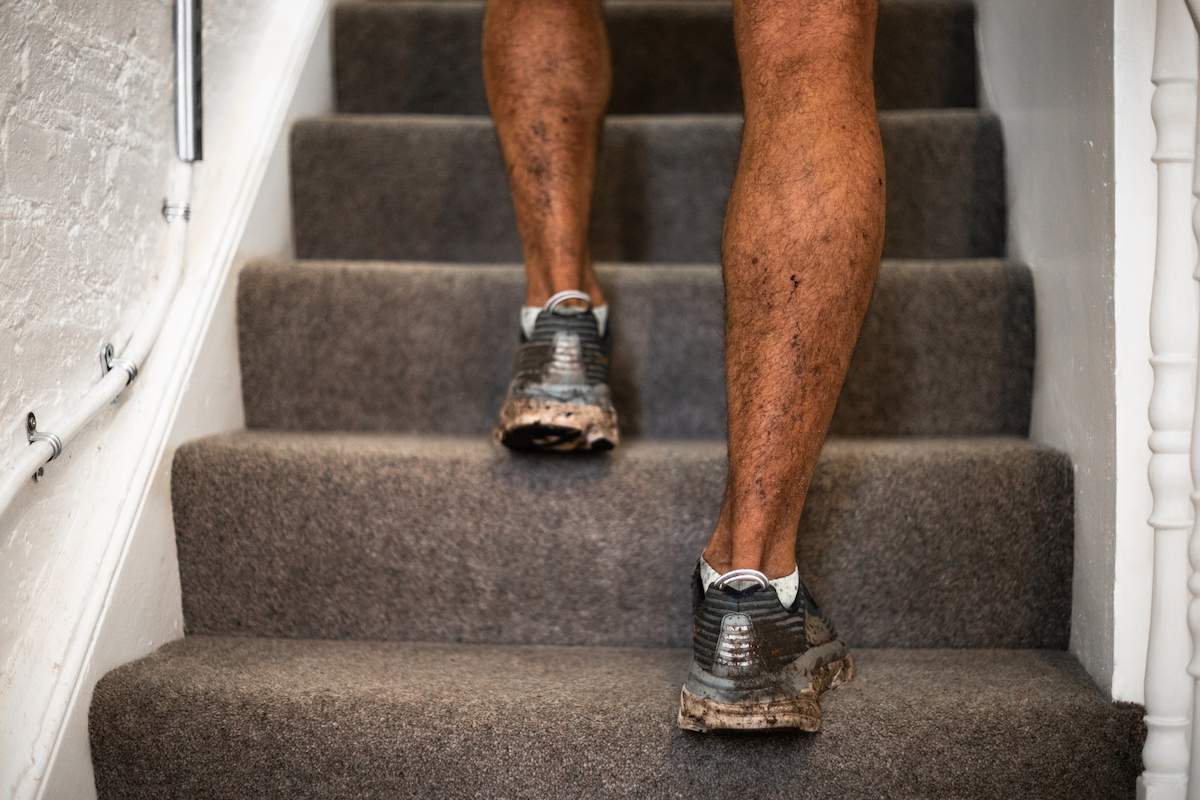 A person wearing muddy running shoes is walking up a grey carpeted staircase.