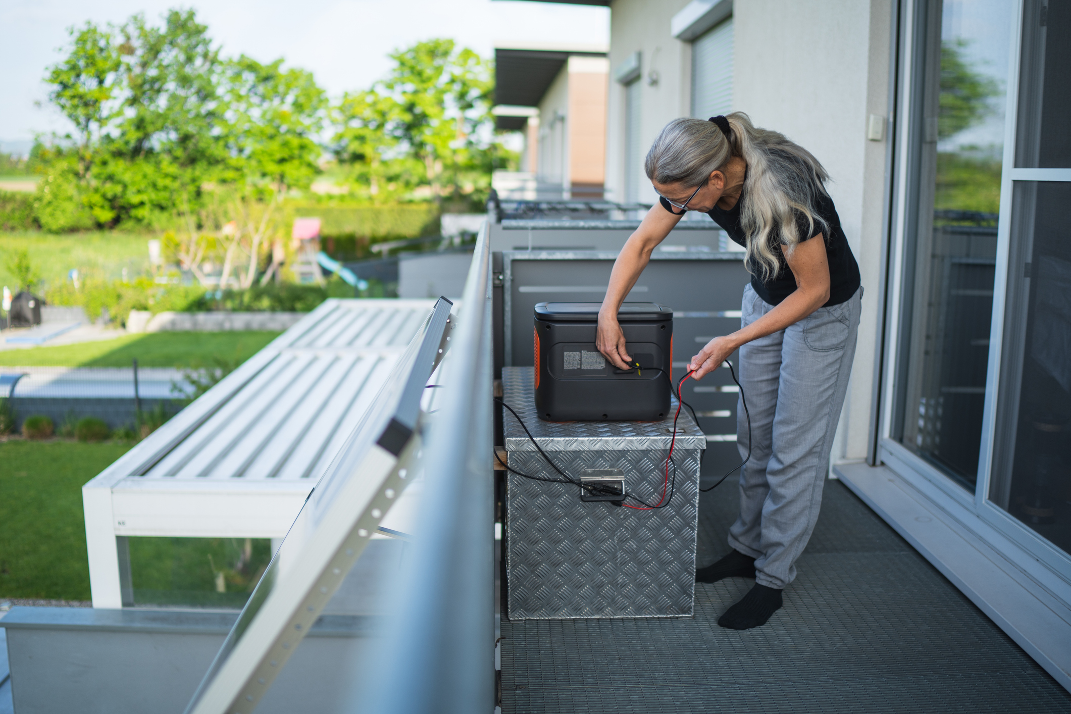 Older woman plugging in cable of solar panel into portable generator on balcony of her house.