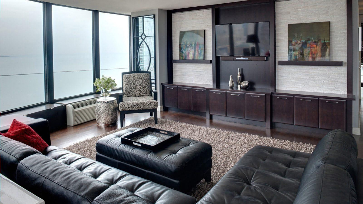 An upscale living room with large glass windows, a long entertainment center, and a large black leather sectional sofa.