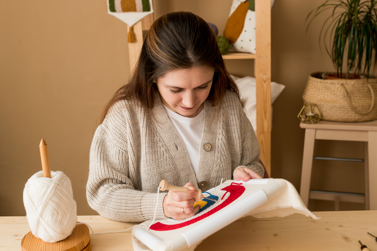 iStock-1395018063 decorate with crafts Female embroidering handmade picture rainbow on canvas. Young woman pushing the punchneedle straight down into the foundation fabric in workshop.