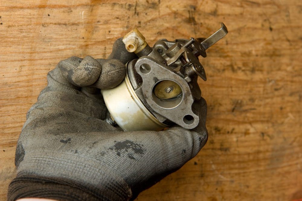 A gloved hand is holding a dirty small engine carburetor against a wooden background.