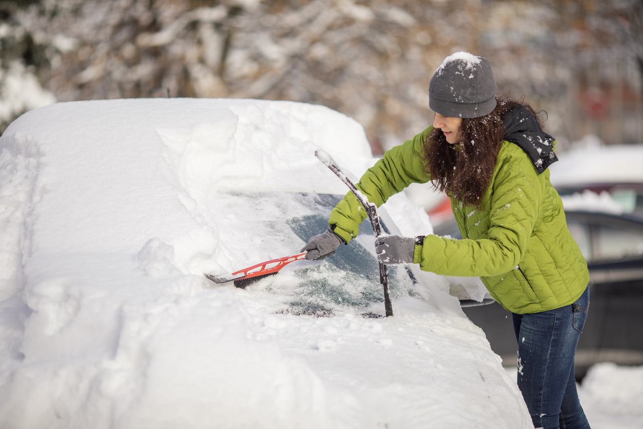 Une femme d'âge mûr nettoie sa voiture de la neige sur le parking. Elle porte des vêtements chauds et utilise une brosse à neige