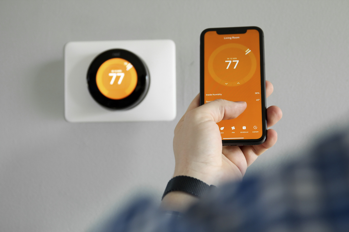 Une personne utilise une application de thermostat orange sur son smartphone pour régler le thermostat intelligent à 77 degrés Fahrenheit.