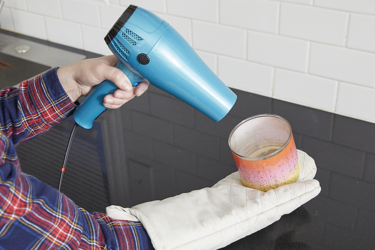 Woman wearing an oven mitt, using a hair dryer to melt candle wax.