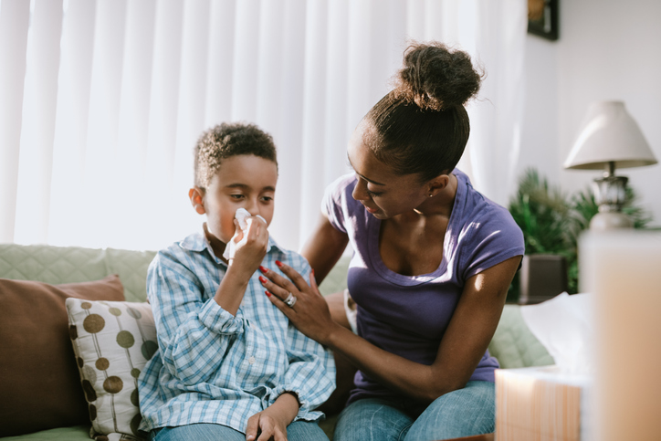 young-mother-comforts-her-son-who-has-a-tissue-to-his-nose-while-they-sit-on-a-tan-sofa-with-a-lamp-in-the-background