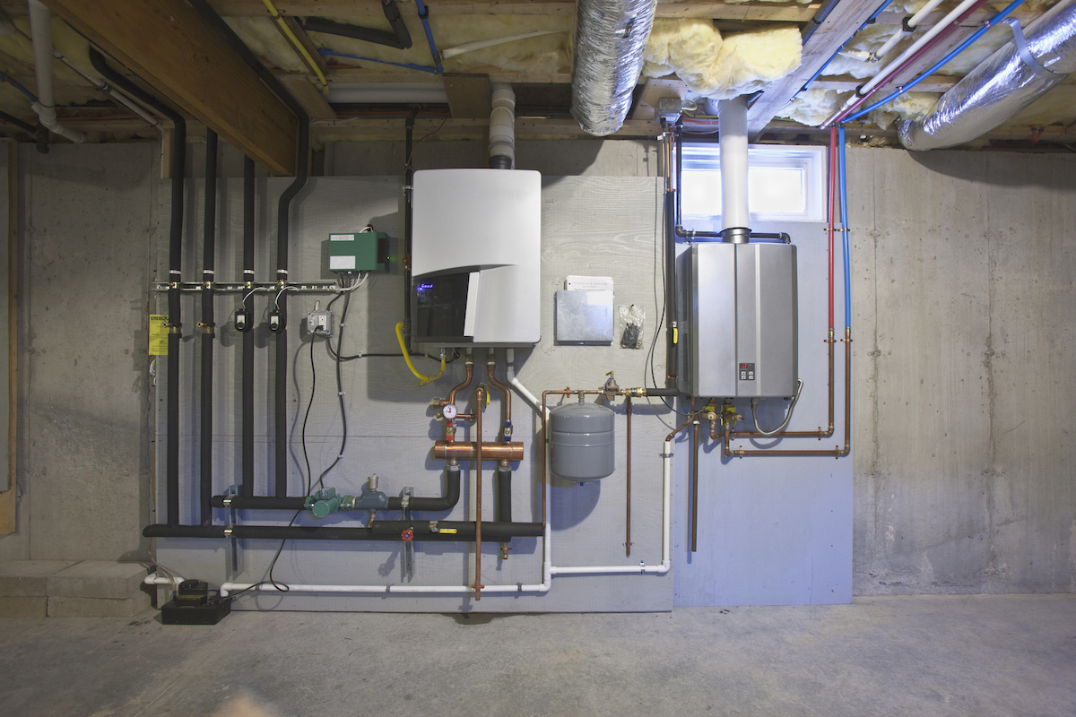 A tankless water heater system is on the wall of a basement underneath a small window.