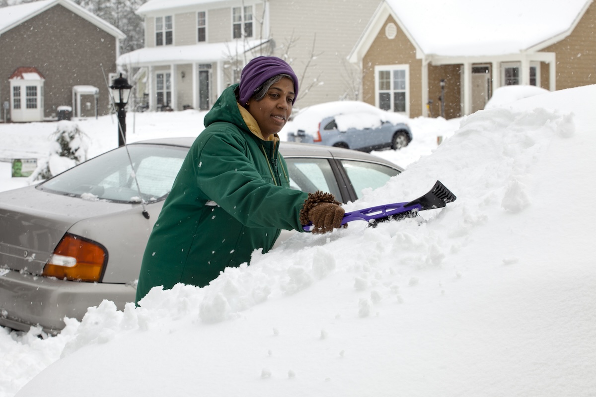 A person dressed in a winter coat, hat, and gloves scraping snow and ice off of a vehicle.