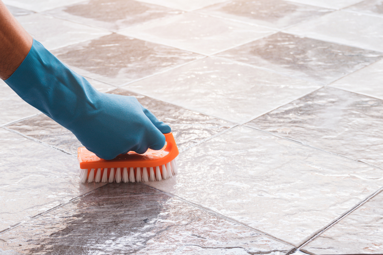 Une personne portant des gants en latex bleu frotte le coulis entre les carreaux de sol avec une brosse à poils durs.