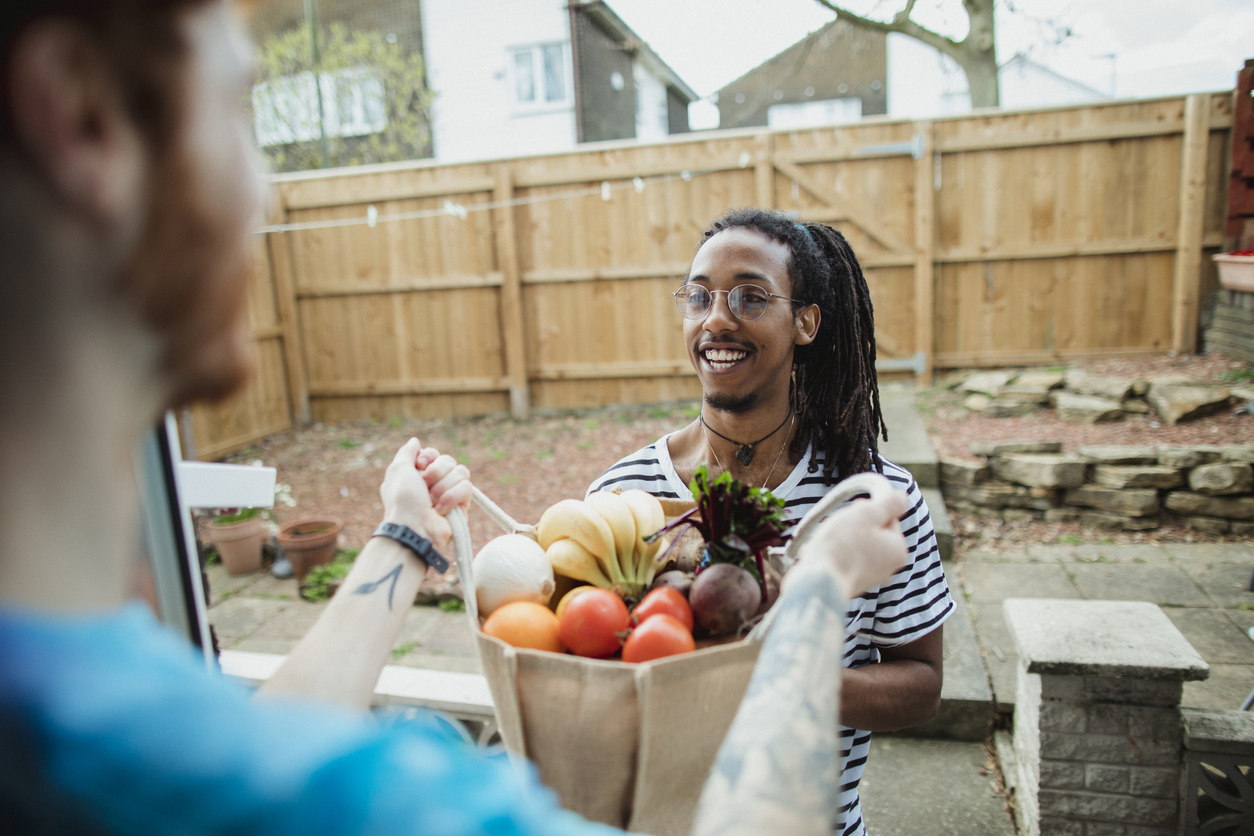 An over the shoulder shot of a young caucasian man holding a large bag of fruit and vegetables, his friend is smiling towards him.