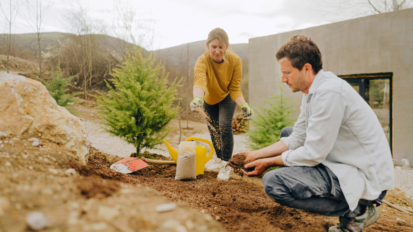 Young couple wearing clothing appropriate for the outdoors while sprinkling grass seed in yard f modern home.