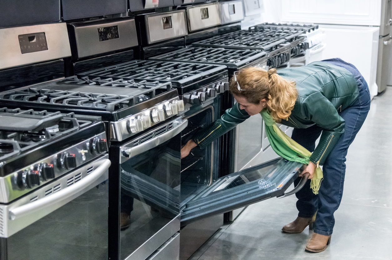 Woman looks inside an oven while shopping an aisle of ovens at a store.