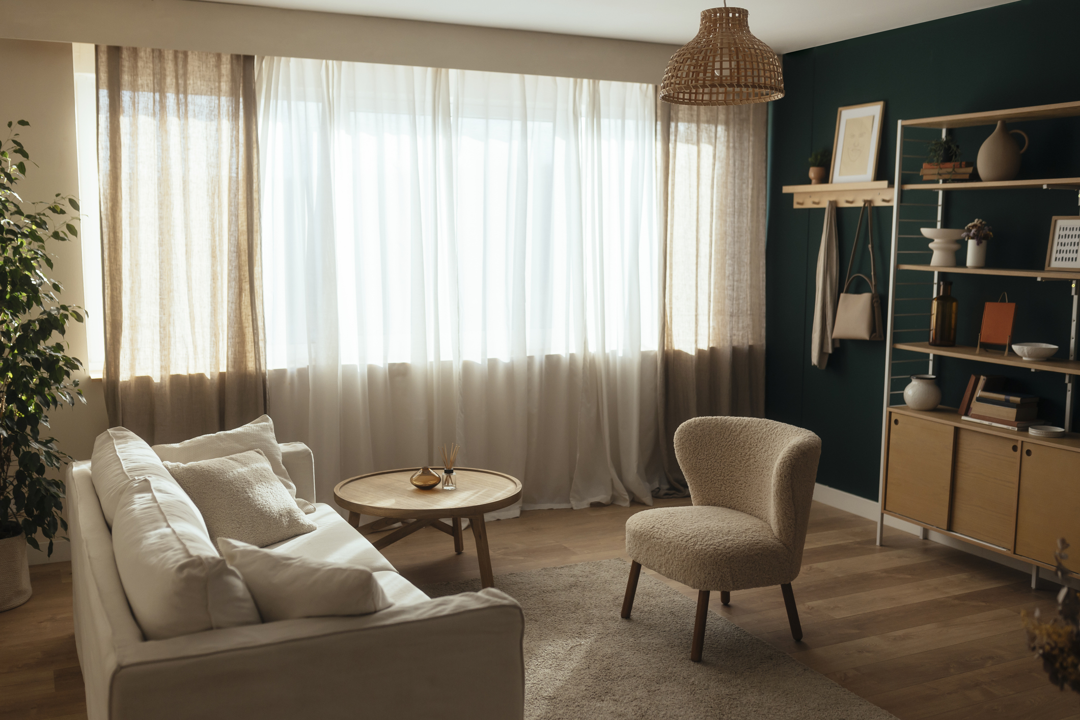 A small living room featuring a white sofa, cream chair, and trendy bookcase is lit only by natural light filtering through shear window curtains.