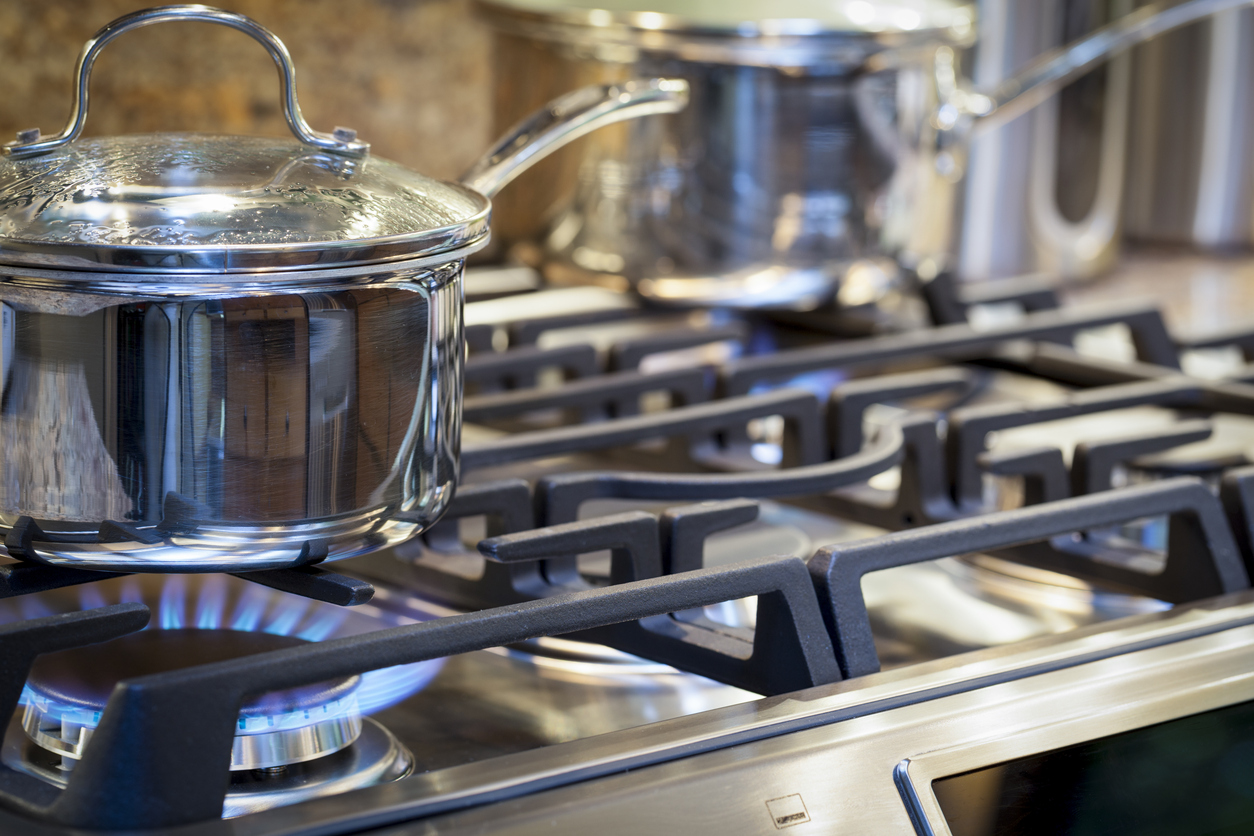 gros plan d'une casserole en acier inoxydable avec poignée et couvercle en verre sur une cuisinière avec du gaz allumé et une autre casserole floue en arrière-plan