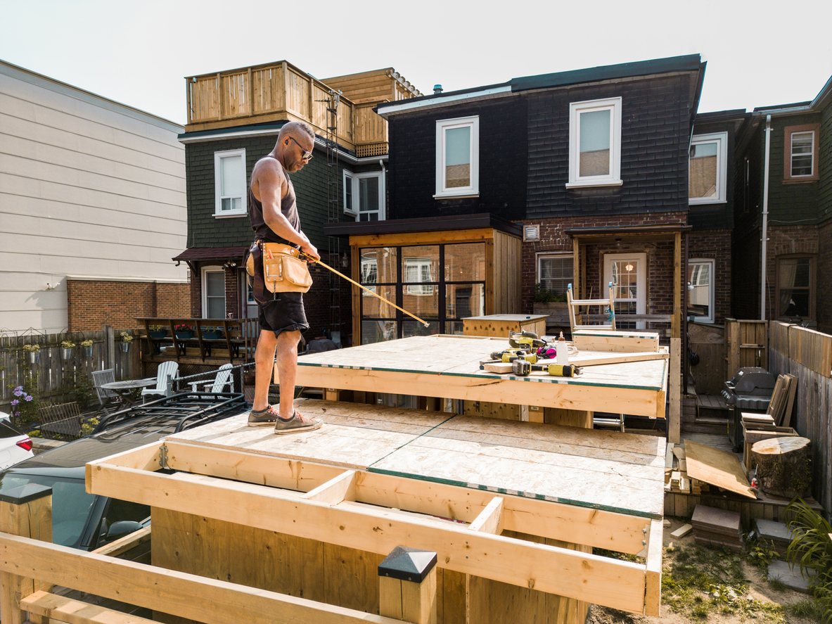 a-workman-measures-for-a-partially-constructed-deck-behind-a-house-with-black-siding