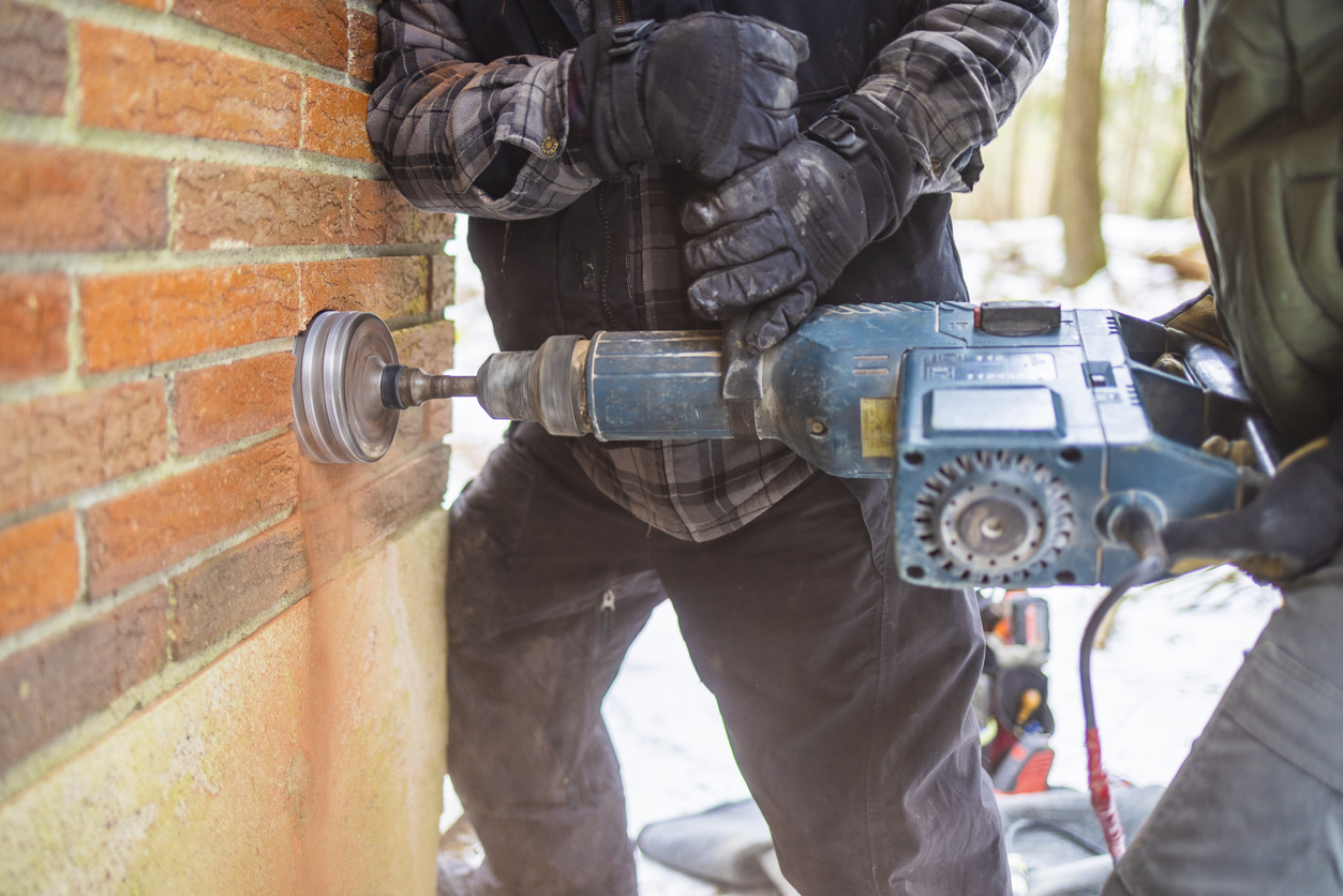 The process of installation radon mitigation system. Two workers are drilling the red brick house wall.