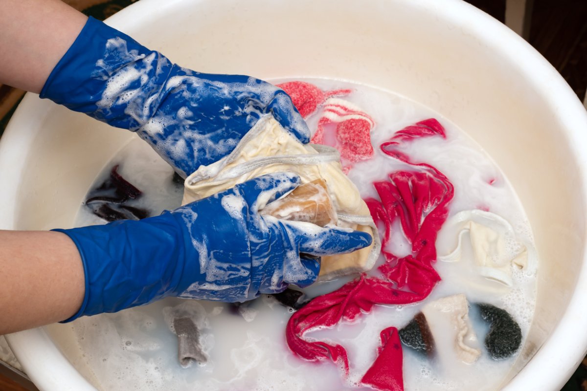 Une personne portant des gants bleus frotte un vêtement tandis que d'autres vêtements trempent dans une bassine savonneuse.