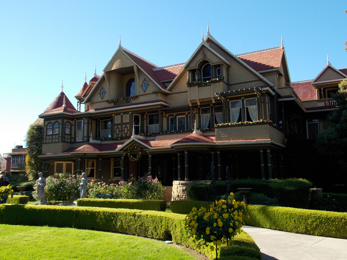 une maison victorienne avec une variété de pignons et de tuiles rouges sur un extérieur beige se trouve derrière des jardins formels contre un ciel bleu