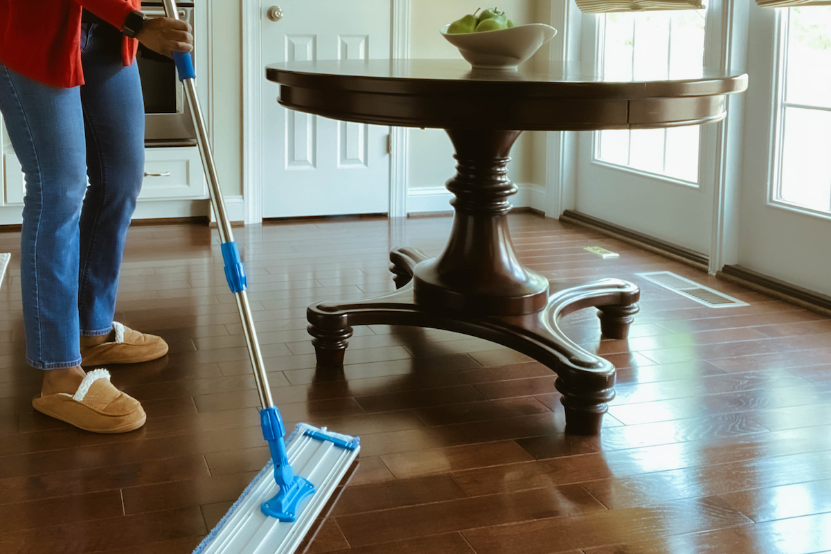 Une personne utilise une vadrouille sèche pour nettoyer le plancher de bois franc autour d’une table en bois.