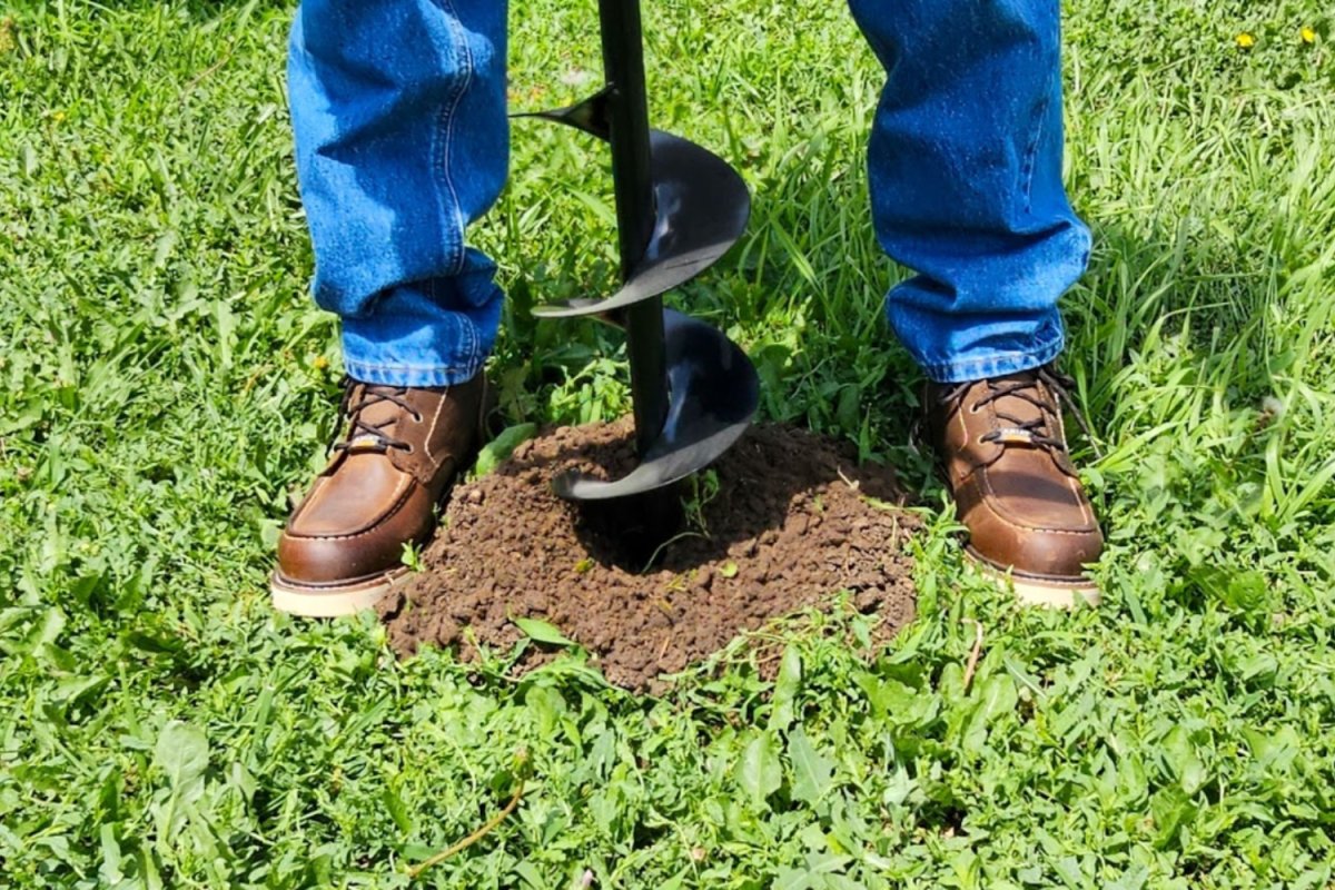 Person in brown work boots and jeans pushing a steel auger into dirt