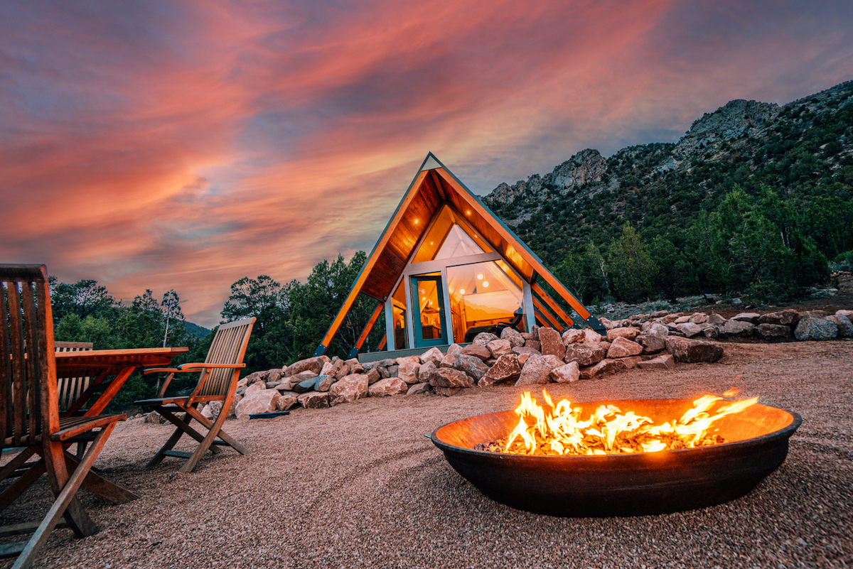 A burning fire pit on gravel lawn is in front of an A-frame house and stunning sunset in background.